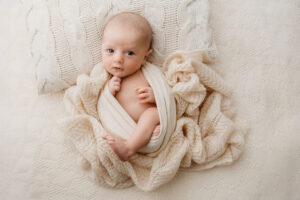 Wide awake newborn baby looking directly at the camera. Baby is holding his hand to his chin. He is cosy and comfortable in a cream shawl. Photographed by Bristol newborn photographer Melanie East