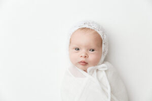 photography of newborn baby looking at camera.  Baby is wearing a white bonnet.  Photographed by Melanie East Photography