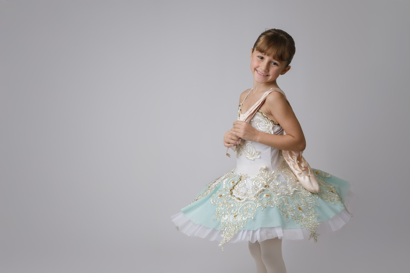 small ballerina wearing a turquoise white and gold tutu holding ballet shoes photographed by Melanie East Dance Photographer in Bristol