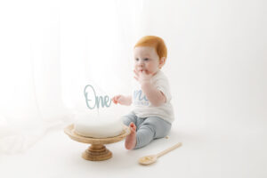 a colour photograph of a one year old baby during a baby's first birthday photo shoot. Baby is wearing a white top and blue trousers.  The cake is white and is on a wooden cake stand and has the word ONE on it in blue. 