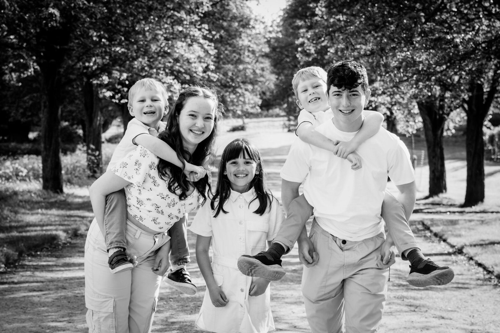 Black and white photograph of two teenagers and three younger children smiling at the camera. There is a background of trees. the image is in black and white. Photographed by Melanie East Family Photographer in Bristol