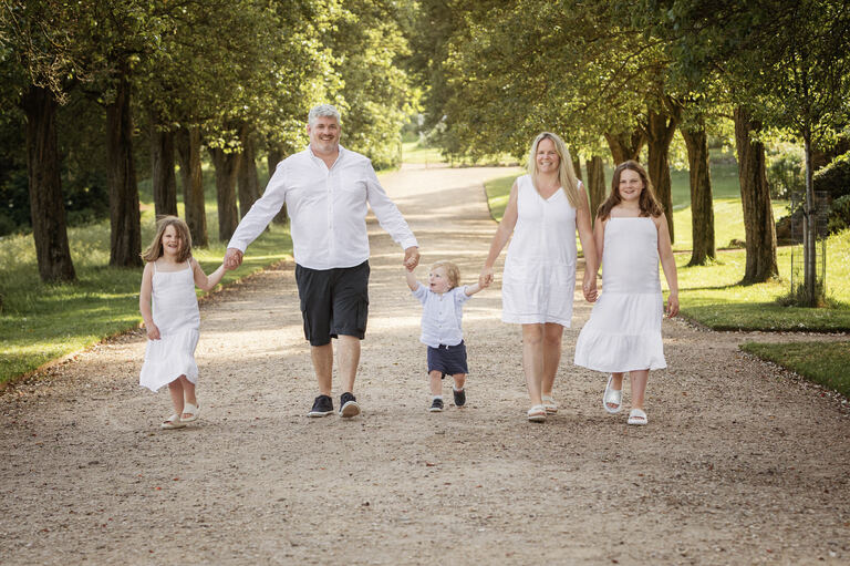 A family of two adults and two children walking towards the camera smiling in a wooden setting. The sun is shining.